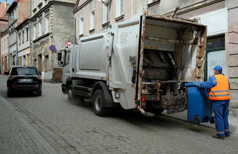 A large white waste collection truck with visible signs of rust and wear at the rear loading area is stationed on a narrow urban street, positioned parallel to a row of older, multi-story buildings with stone and brick facades. A worker in a blue uniform with an orange high-visibility vest and matching blue cap is standing at the back of the truck, emptying a blue wheeled refuse bin into the hopper. The street surface is cobblestone, with a slight incline, and a black car is parked nearby on the left side of the street. Behind the truck, there are two traffic restriction signs mounted on a metal pole, indicating parking limitations. The surrounding environment shows a mix of architectural details, including large windows and weathered exteriors, suggesting a historic or densely populated area. The scene is lit naturally, with diffuse daylight, emphasizing the functional nature of waste collection, which is part of private or independent rubbish removal services, supporting the broader context of alternative waste management solutions outside official municipal collection. Waste Removal West Hampstead is involved in responsible rubbish clearing operations here.