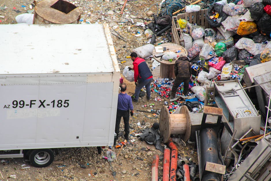An aerial view of a large outdoor rubbish pile on a dirt surface with scattered debris, plastic bottles, paper, and discarded materials. Several black, white, and multi-colored rubbish bags are piled haphazardly on the right side, some partially torn open. In front of the rubbish, two individuals are sorting through the debris; one wears a red jacket and dark pants, while the other is in darker clothing and bent over. To the left, a white large box truck with the identifier 