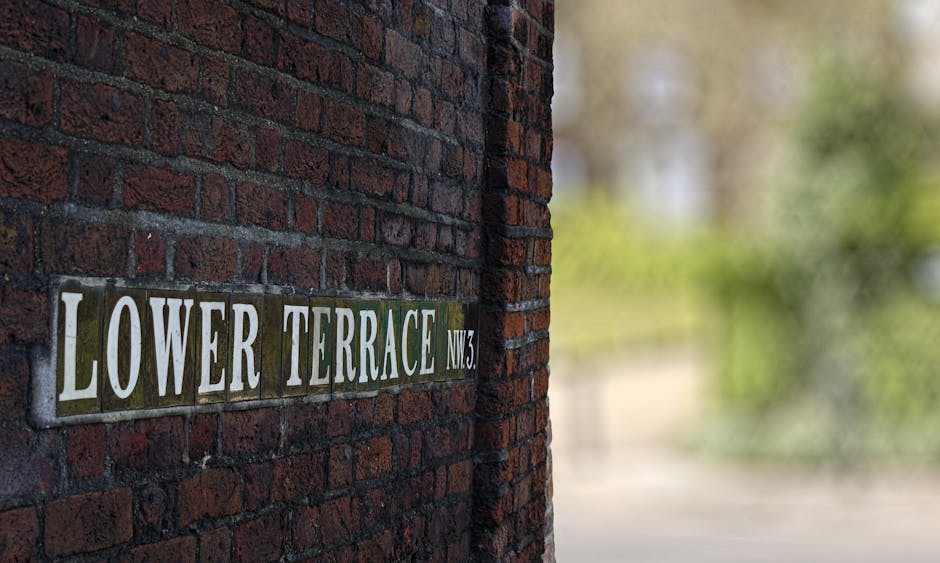 A close-up view of a brick wall displaying a metal street sign that reads 'Lower Terrace NW3' with white and dark green tiles. The sign is attached to a red-brown brick surface with a textured finish. In the background, there's a blurred outdoor scene featuring greenery and foliage, indicating a residential or urban setting. The lighting appears natural, with daylight illuminating the bricks and the sign, providing clear visibility of the materials and textures. This scene exemplifies a typical exterior setting where waste removal or rubbish collection might be arranged privately, such as in a residential area serviced by independent waste management providers like Waste Removal West Hampstead. The scene's composition and environment support the context of localized rubbish handling, highlighting the importance of on-site clearance solutions for properties in the region.