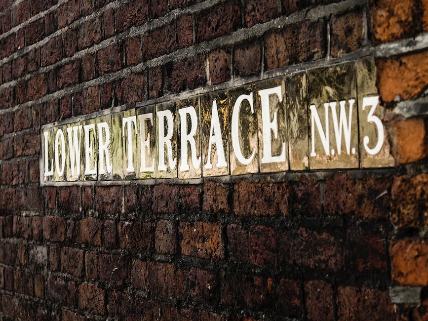 A close-up view of a brick wall displaying a metal street sign that reads 'Lower Terrace NW3' with white and dark green tiles. The sign is attached to a red-brown brick surface with a textured finish. In the background, there's a blurred outdoor scene featuring greenery and foliage, indicating a residential or urban setting. The lighting appears natural, with daylight illuminating the bricks and the sign, providing clear visibility of the materials and textures. This scene exemplifies a typical exterior setting where waste removal or rubbish collection might be arranged privately, such as in a residential area serviced by independent waste management providers like Waste Removal West Hampstead. The scene's composition and environment support the context of localized rubbish handling, highlighting the importance of on-site clearance solutions for properties in the region.