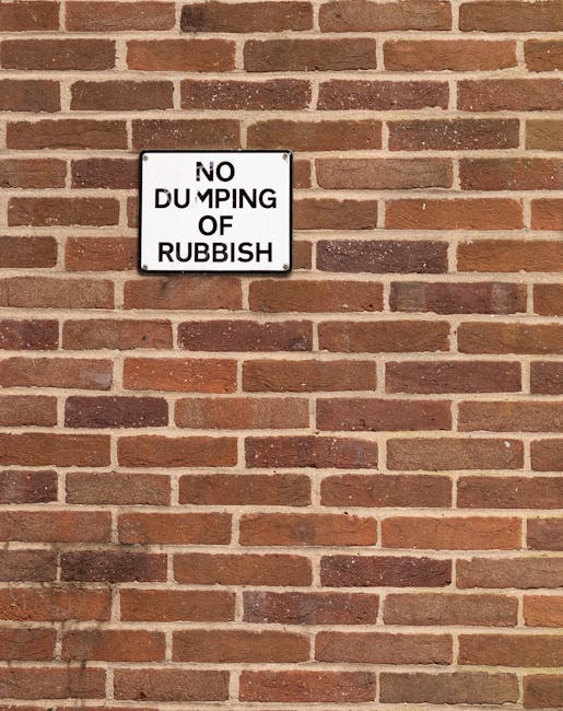 A square black and white metal sign mounted on a red brick wall displaying the message 'NO DUMPING OF RUBBISH' in bold black text, with the words arranged in four lines. The brick wall features horizontal rows of uniformly sized bricks with varying shades of reddish-brown, beige, and dark brown, connected by light mortar joints. The sign is positioned towards the upper left portion of the image, with the bricks extending across the entire frame, creating a textured and durable backdrop typical of urban environments where waste management and rubbish removal services are often required. The lighting is even, highlighting the natural texture of the bricks and the clear, utilitarian design of the sign, which serves as a visual deterrent against unwanted waste disposal in this area managed by waste removal services like Waste Removal West Hampstead.