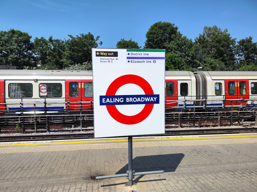 A clear view of a station sign reading 'Ealing Broadway' with a red circular London Underground logo and a blue strip across the middle, displaying the station name in white. Behind the sign, there is a modern train with a silver body and red and blue accents parked on the railway tracks. The train's windows have reflective glass, and some interior elements are faintly visible. The surrounding environment includes green leafy trees against a bright blue sky, indicating a sunny day. The station platform has a textured, paved surface with yellow safety lines along the edge, and a metal fence separates the platform from the train tracks. This scene represents a typical urban railway station with a focus on public transportation infrastructure, related to efficient on-site or alternative transportation methods, complementing the context of waste management services by emphasizing community mobility infrastructure.