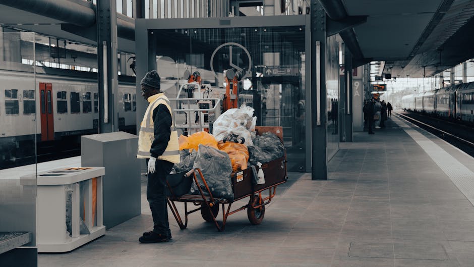 At a modern, open-air railway station platform during daylight hours, a worker dressed in dark clothing, a grey beanie, and a high-visibility vest stands next to a red metal-wheeled trolley filled with various bags and discarded items, including black, grey, and orange plastic bags, some appearing partially filled and loosely tied. The trolley is positioned close to the edge of the platform, which has a textured grey surface and a designated safety zone marked by a tactile paving strip along the track. Behind the worker, there is a glass-enclosed lift shaft with metallic framing, and visible machinery or infrastructure components inside. To the left, a row of vintage-style train carriages with white bodies and red doors is parked parallel to the platform. On the right, the platform extends into the distance where a few other passengers are visible and the train tracks continue straight ahead. The overall scene suggests a setting where waste or rubbish removal services, such as those provided by Waste Removal West Hampstead, are involved in clearing discarded items directly from public transport areas, supporting alternative waste handling outside standard station waste collection facilities.