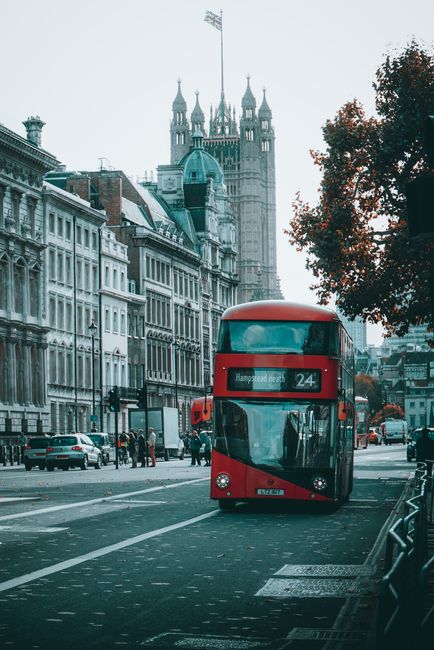 A red double-decker bus traveling along a city street with historic buildings and a prominent clock tower visible in the background. The bus displays the route number 24 and the destination, 'Hampstead Heath,' on the electronic sign at the front. To the right, there are parked cars and a sidewalk with pedestrians walking. The scene is set during daytime with natural lighting, and the environment suggests an urban area with a mix of modern transportation and historic architecture. The street is lined with a few trees, one of which is partially visible on the right, providing some greenery to the scene. The overall atmosphere is typical of central London, with the bus as the focal point, emphasizing city transport and urban life, which relates to independent transport and waste removal services in busy city environments managed by companies like Waste Removal West Hampstead.