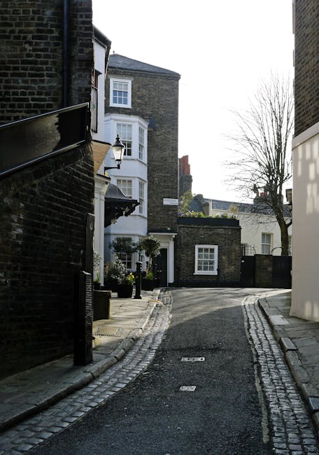 The image depicts a narrow cobbled alleyway in a residential area, flanked by dark brick buildings on both sides. On the left, a black-painted metal railing runs along the edge of a building, with a traditional-style street lamp mounted on a white column near the corner. Adjacent to this building, potted plants with greenery are placed near the entrance, which features a black door. The buildings display classic Victorian or Georgian architectural elements, including bay windows with white frames and decorative external finishes. The alleyway curves gently to the right towards a small square or junction, with a small brick structure visible in the middle distance. In the background, there is a leafless tree with bare branches, indicating late autumn or winter. The scene is illuminated by daylight, with a clear sky overhead. The overall setting suggests a quiet, historic part of town that may require occasional private waste removal, aligning with the services of Waste Removal West Hampstead providing alternative rubbish clearance solutions for residents in the area interested in independent collection or on-site clearance options.