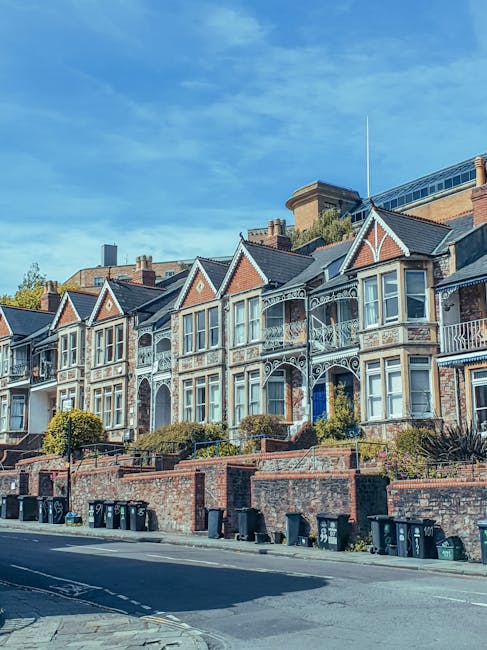 A row of Victorian-style terraced houses with ornate white decorative ironwork on the balconies and bay windows, featuring red brick facades and grey slate roofs, situated along a paved street with a brick wall and multiple black and green wheelie bins lined up at the curb. The houses are set on a slightly elevated ground, with small landscaped front gardens and low brick walls, under a clear blue sky with a few wispy clouds. The scene depicts an urban residential area in West Hampstead where private rubbish collection services might be employed for waste removal, as suggested by the presence of various bins positioned outside the properties. The overall setting emphasizes traditional architecture combined with modern waste management infrastructure, consistent with local waste collection practices in the UK.
