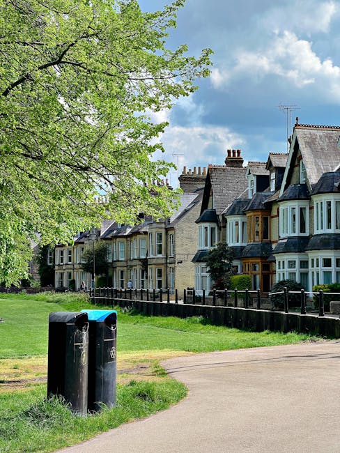 A row of Victorian-style terraced houses with ornate white decorative ironwork on the balconies and bay windows, featuring red brick facades and grey slate roofs, situated along a paved street with a brick wall and multiple black and green wheelie bins lined up at the curb. The houses are set on a slightly elevated ground, with small landscaped front gardens and low brick walls, under a clear blue sky with a few wispy clouds. The scene depicts an urban residential area in West Hampstead where private rubbish collection services might be employed for waste removal, as suggested by the presence of various bins positioned outside the properties. The overall setting emphasizes traditional architecture combined with modern waste management infrastructure, consistent with local waste collection practices in the UK.