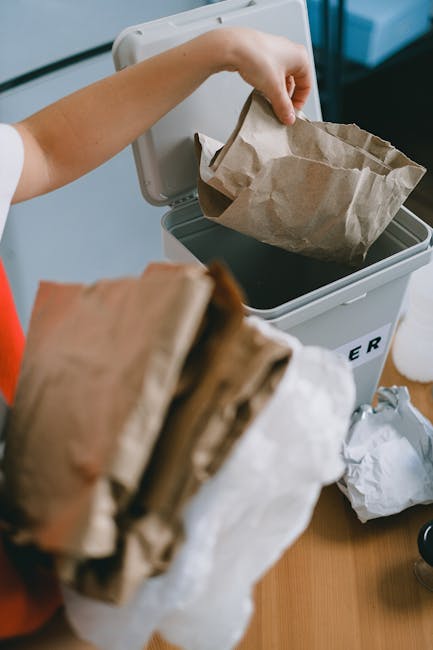 A person placing a crumpled brown paper bag into a white recycling bin with a black lid, situated indoors on a wooden surface. The individual's arm and hand are visible, with the hand gripping the paper bag, which shows creases and folds. In the foreground, additional crumpled paper bags are held, partially obscuring the view of the bin. The environment appears to be a domestic or small office setting, with a focus on waste separation and disposal. The recycling bin is designed for cardboard and paper waste, supported by Waste Removal West Hampstead's focus on alternative waste handling options. The scene emphasizes proper rubbish sorting and the use of dedicated containers for recycling paper products, aligning with the service of managing waste that might otherwise be subject to local authority collection restrictions or private disposal methods.